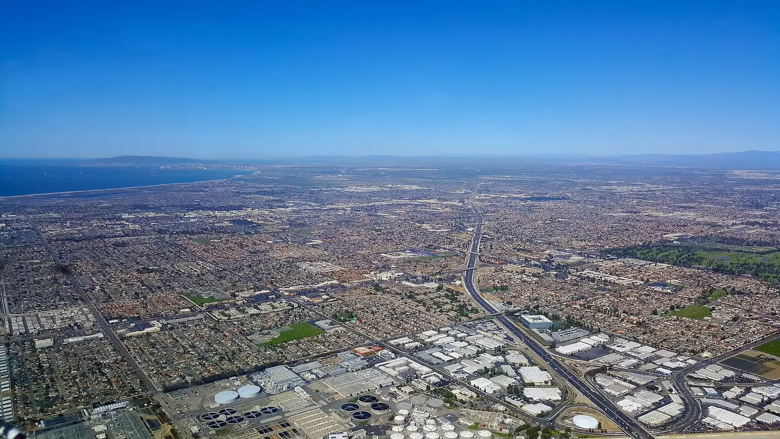 Aerial view across inland Huntington Beach and Fountain Valley, showing the flat grid of neighborhoods, the 405 freeway cutting through, and the ocean along the far horizon.